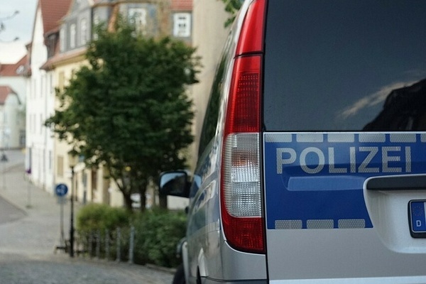 Auf dem Bildausschnitt ist die Rckseite eines Polizeiautos und der Schriftzug Polizei in Grau auf blauem Untergrund. In der Ferne sieht man verschwommen eine Strae und einen Baum sowie eine Huserreihe.